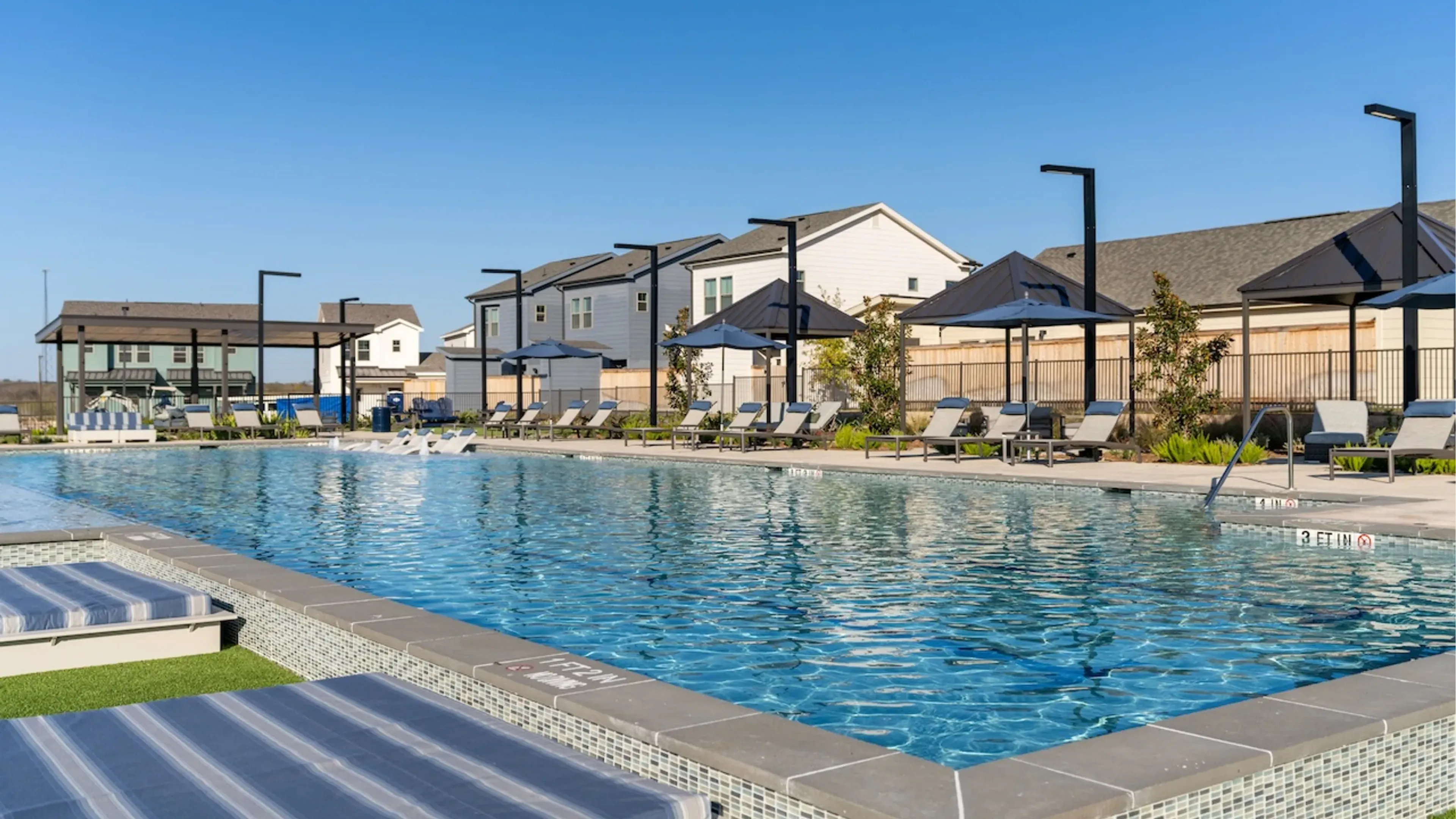 Outdoor swimming pool with lounge chairs and umbrellas on a sunny day, surrounded by residential buildings in the background
